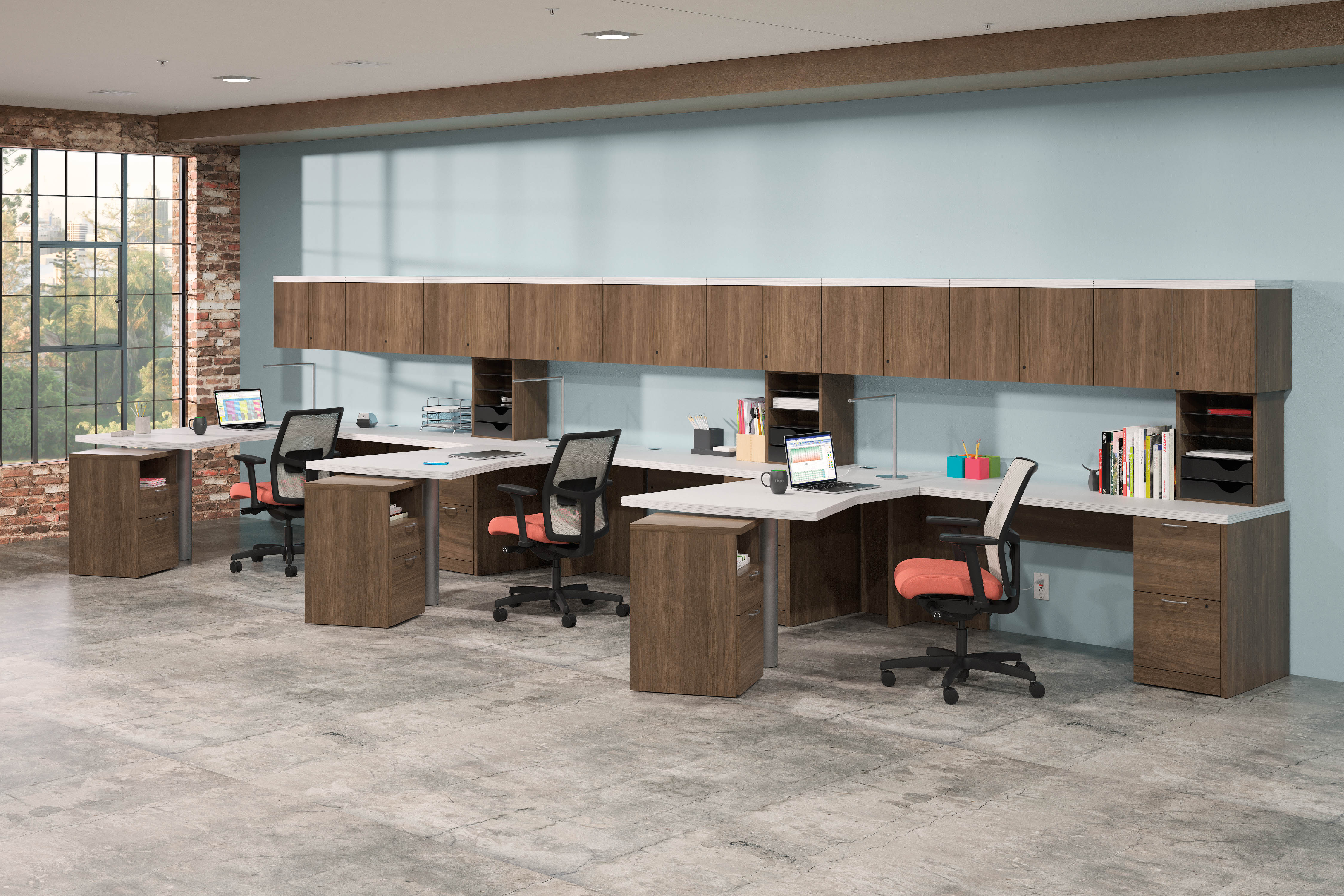 HON Valido desking system of three workstations in an L shape against a wall. Desking system with overhead bins is in a maple colored laminate with white worksurfaces. Each space is complete with a task chair with a red seat and grey mesh backing.