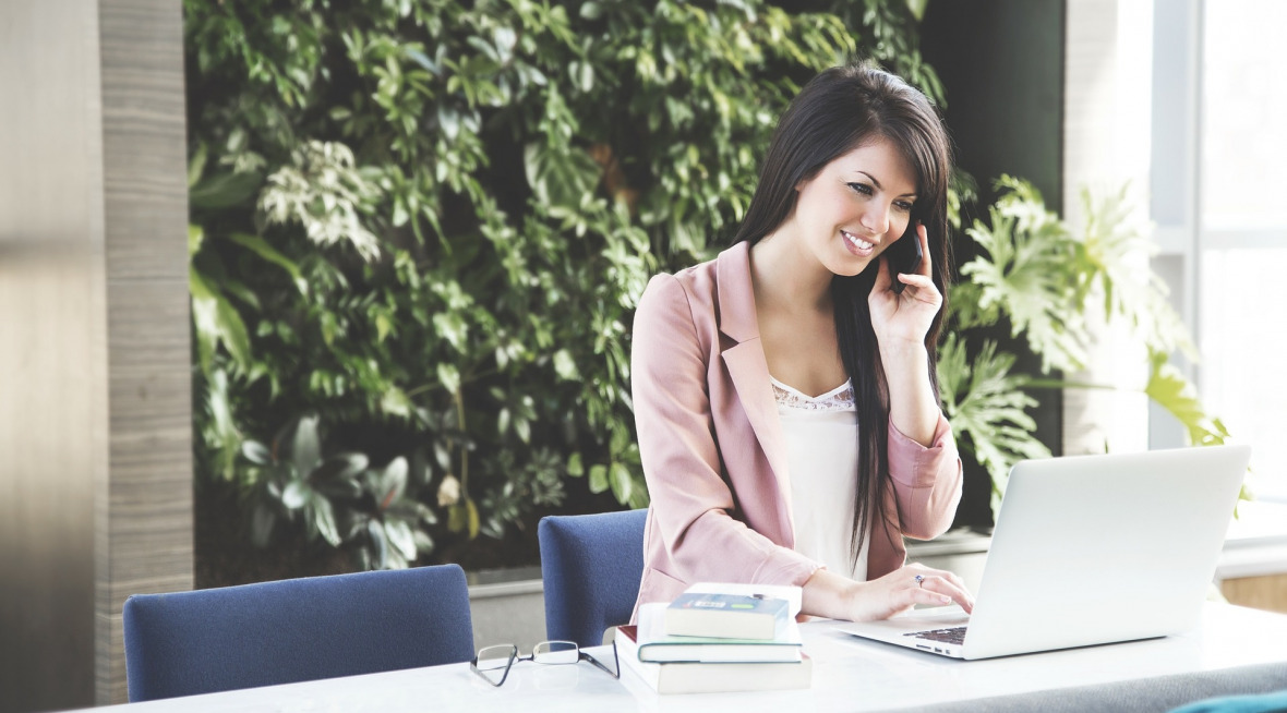 Woman with brown hair holding a cell phone and talking while she is working on a lap top at a table desk. Greenery can be seen in the background of her workspace.