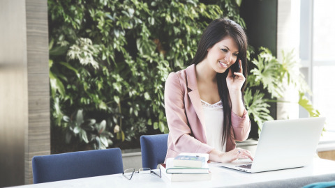 Woman with brown hair holding a cell phone and talking while she is working on a lap top at a table desk. Greenery can be seen in the background of her workspace.