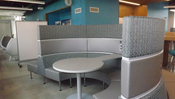 Coalesse lounge seating ganged together and arranged in a half circle with a platinum sold table extending through the middle of the half circle. Grey Steelcase Jenny chairs can be seen in the background