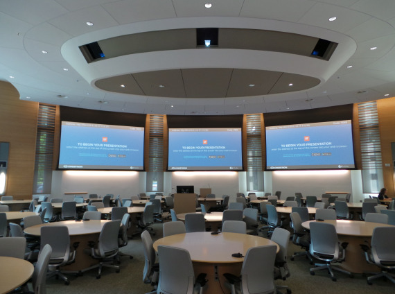 Three monitors on walls in front with multiple tables and chairs in conference extra large conference room with hundreds of task chairs and many blonde colored round conference tables.