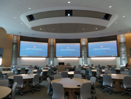 Three monitors on walls in front with multiple tables and chairs in conference extra large conference room with hundreds of task chairs and many blonde colored round conference tables.