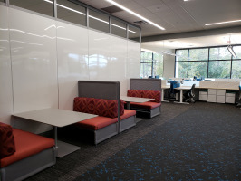 White Steelcase markerboard privacy walls on the left with black Coalesse Lagunitas lounge seating with red cushions. Hotel desking with blue chairs can be seen in the background.