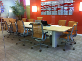 Conference room with orange colored accent wall with a silver art design. Long blonde conference table and ten orange chairs.