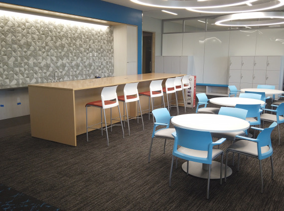 Cafeteria and meeting area with white tables, blue chairs, red stools, and a blonde laminate colored counter space.
