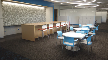 Cafeteria and meeting area with white tables, blue chairs, red stools, and a blonde laminate colored counter space.