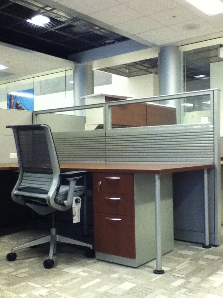 Cubicle desk with glass stackers on top of panels, a cherry colored laminate desk and a Steelcase Leap chair with a file cabinet under the desk.